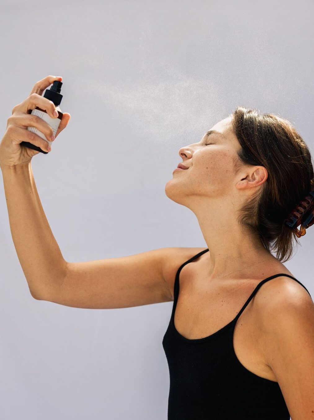 The side profile of a woman with short hair and a black tank top spraying Primally Pure's Everything Spray on her face.