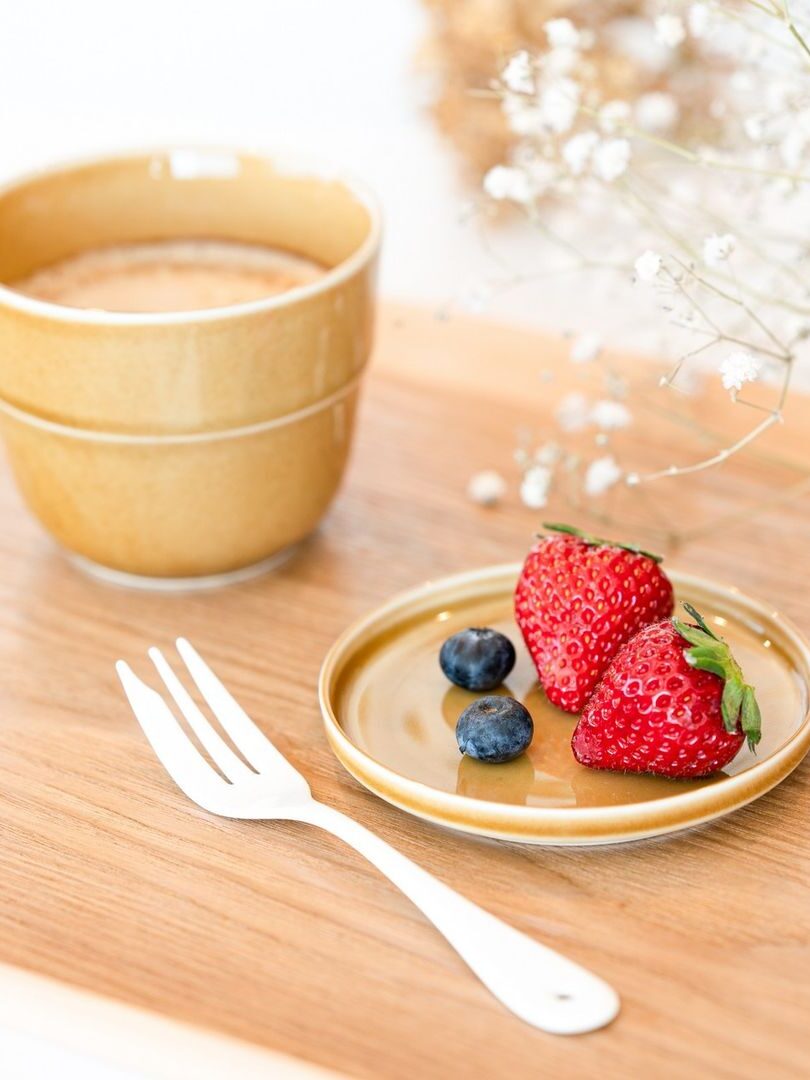 A Tripware ceramic plate in yellow with 2 strawberries and 2 blueberries on the plate. A silver fork is next to the plate while a yellow Tripware ceramic mug is in front of the plate. All items are placed on a wooden tray.