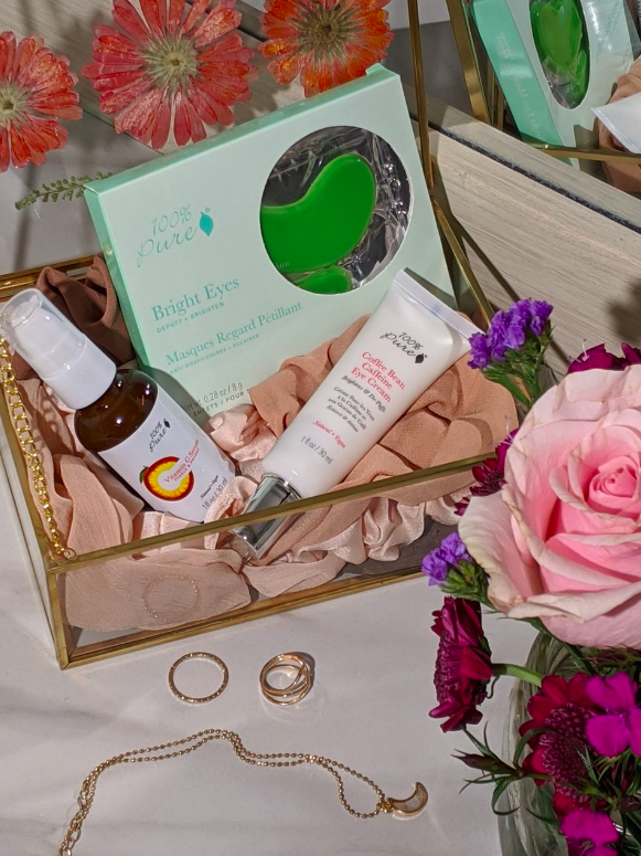 100% Pure's Coffee Bean Caffeine Eye Cream in a clear flip lip, gold rimmed box with other skincare products, flowers, jewelry and pink cloth in and around the box.
