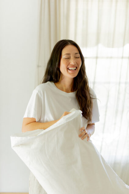 A woman with long dark hair wearing a white t-shirt smiles while holding a large white pillow in a bright room with light curtains.
