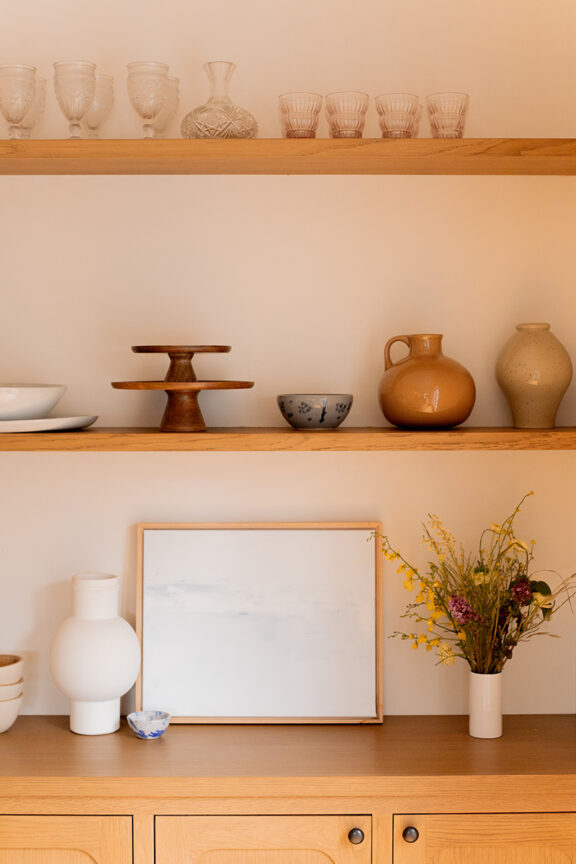 A wooden shelving unit displaying various items including glasses, vases, bowls, and a framed artwork, with a bouquet of wildflowers on the lower shelf.