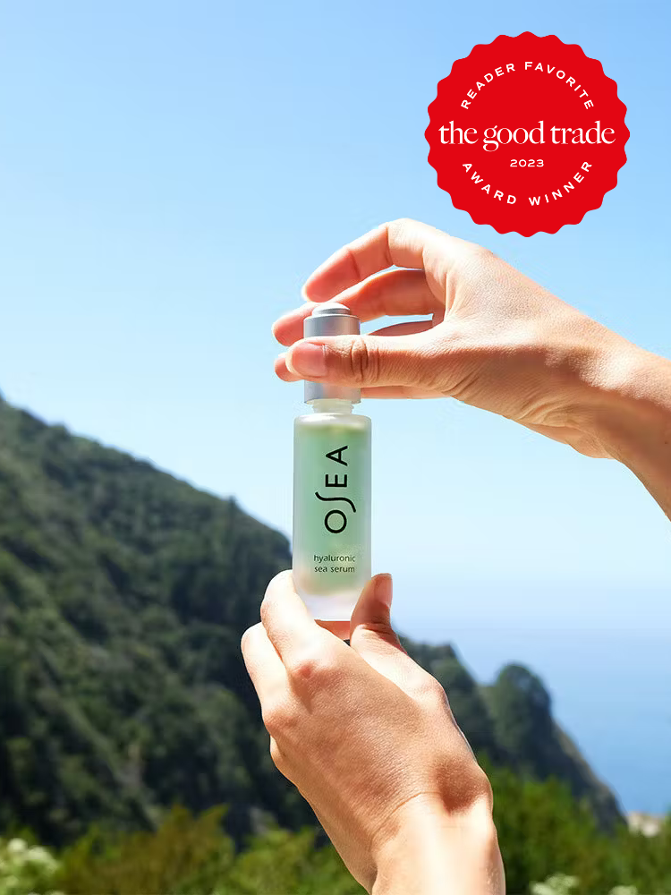 Two hands holding up a bottle of Osea's Hyaluronic Sea Serum outside with a tree covered hill in the distance. The Good Trade's 2023 Reader Award Sticker in red in on the top right corner of the image.
