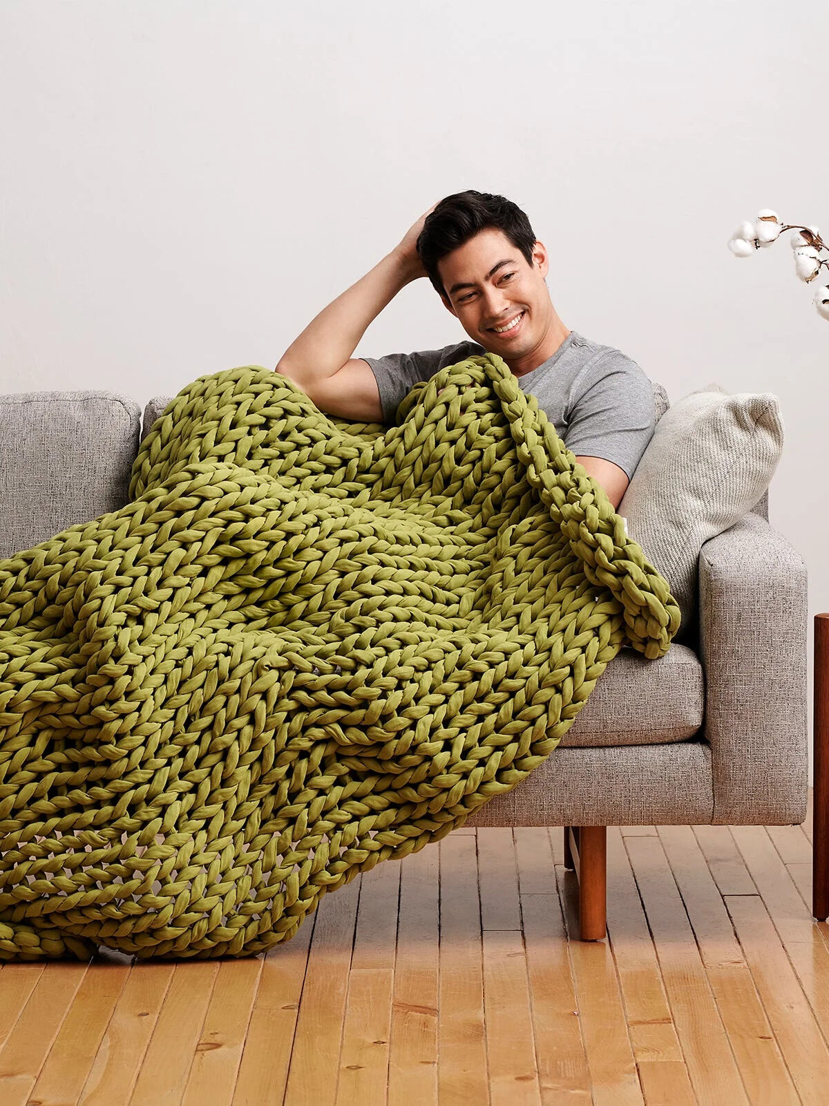 A model laying on a grey sofa with a moss green Bearaby weighted blanket on him. 