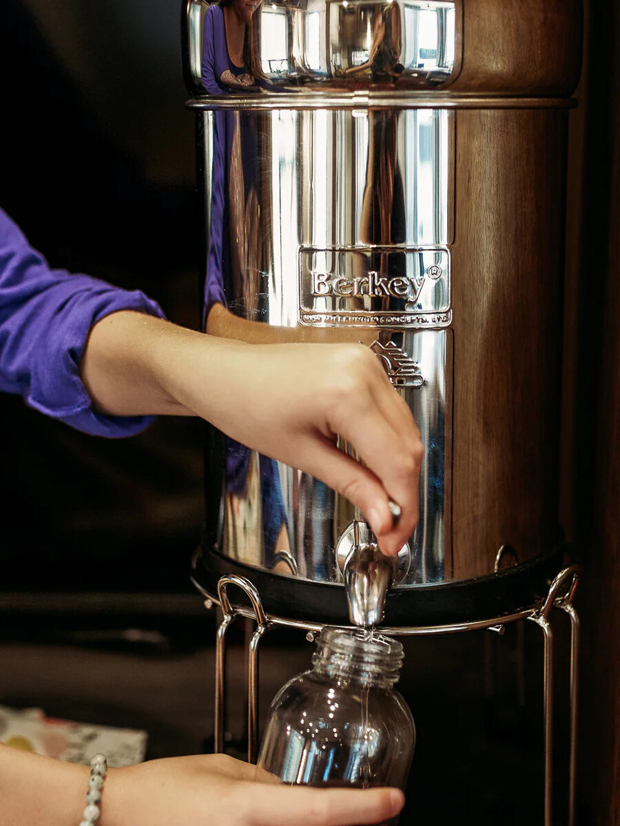 An arm wearing a purple sweater pouring water from the spout of the Berkey Water Filter into a glass bottle.
