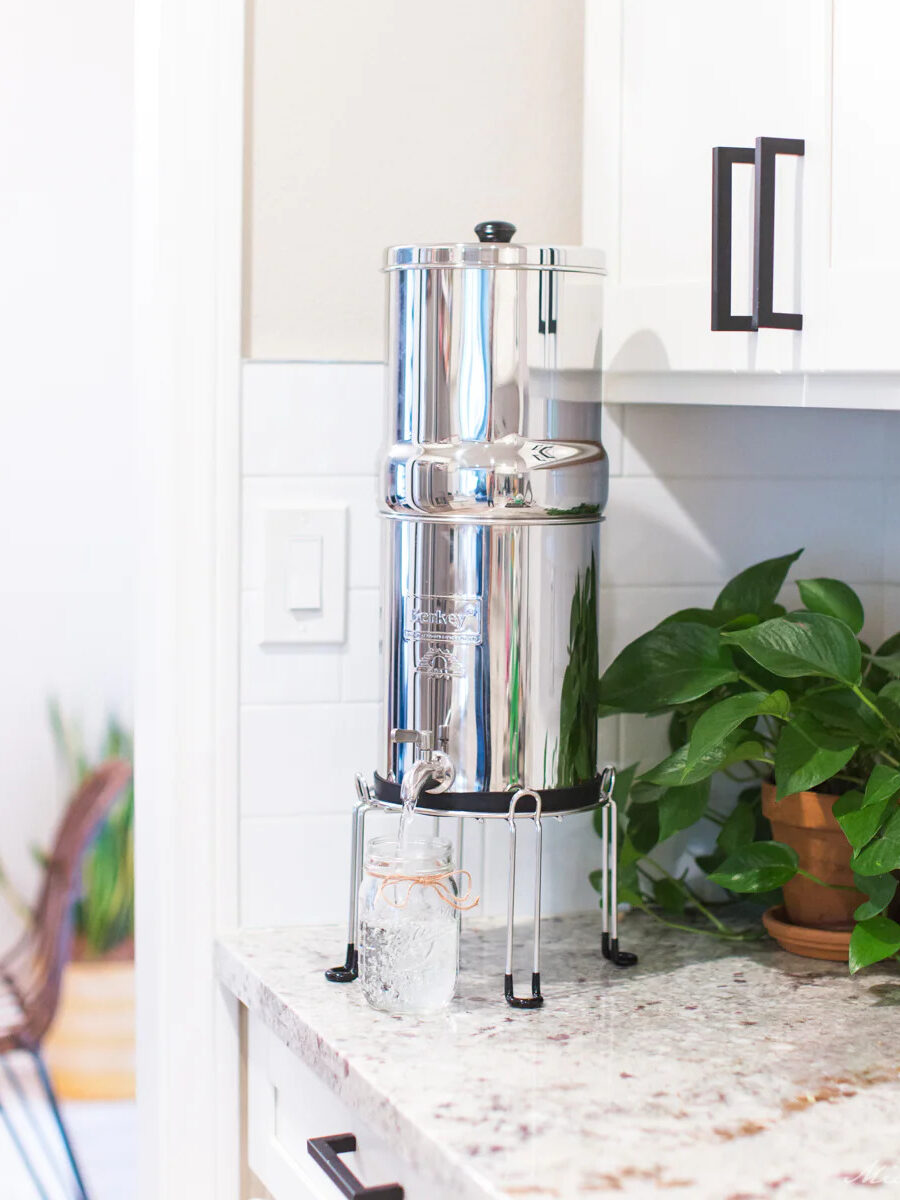 The Berkey Water Filter System set on a kitchen counter with plants behind it and the a dining room chair in the distance.