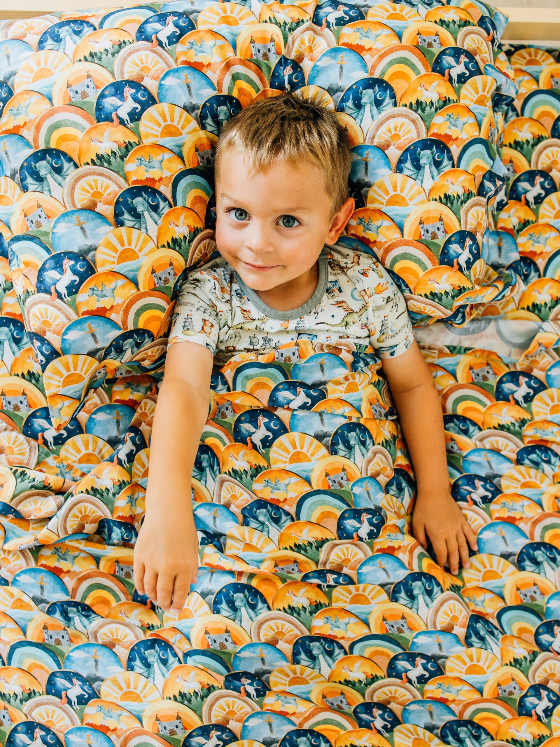 A child laying in a unicorn, castle, dragon, sunshine printed set of sheets in the colors blue and orange. By Brightly.