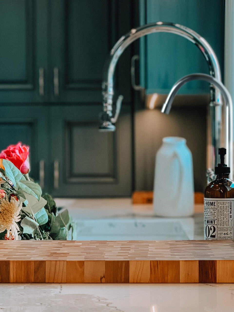 A kitchen sink and spout with a cutting board in front of it and flowers to the left of the spout.