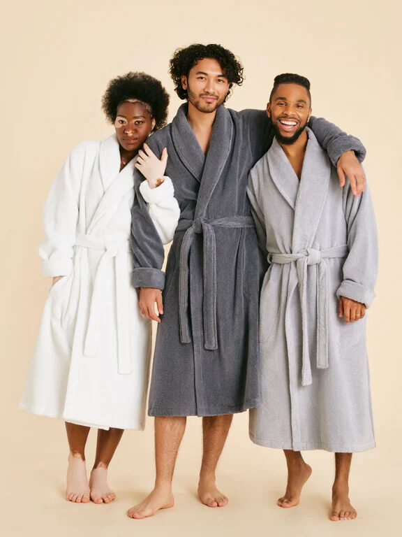 Three models wearing Brooklinen sustainable robes in white, dark grey and light grey.