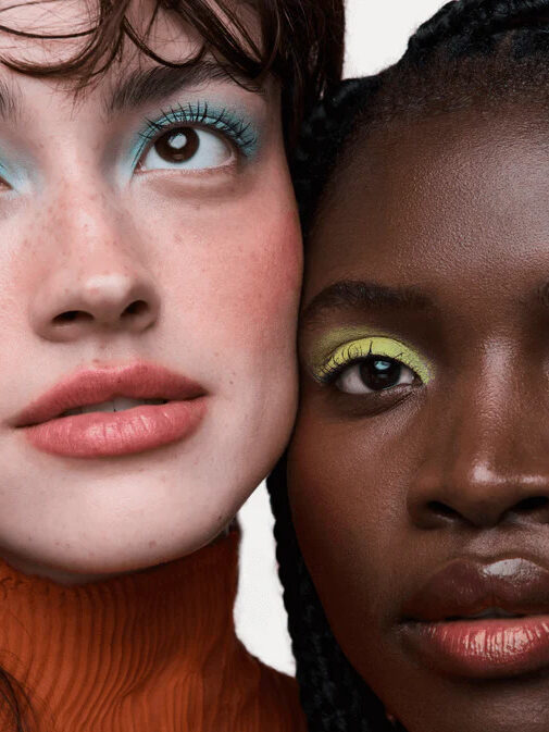 A closeup of two models leaning against each other, one wearing bright blue eyeshadow and the other bright green eyeshadow. By Credo.
