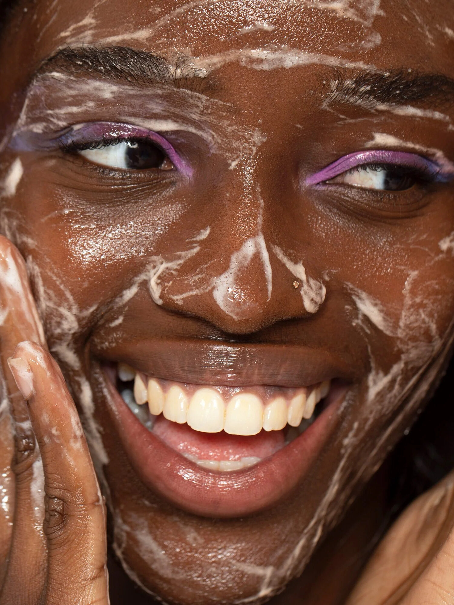 A woman with pink and purple eyeshadow applying a Herbivore cleansing product to her face while smiling.