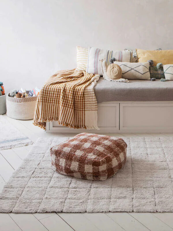A brown checkered pouf by Lorena Canals styled on the floor of a bedroom. 