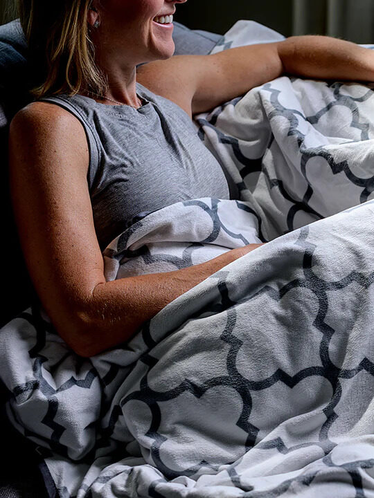 A model laying on a couch with a grey patterned Luxome weighted blanket across her. 