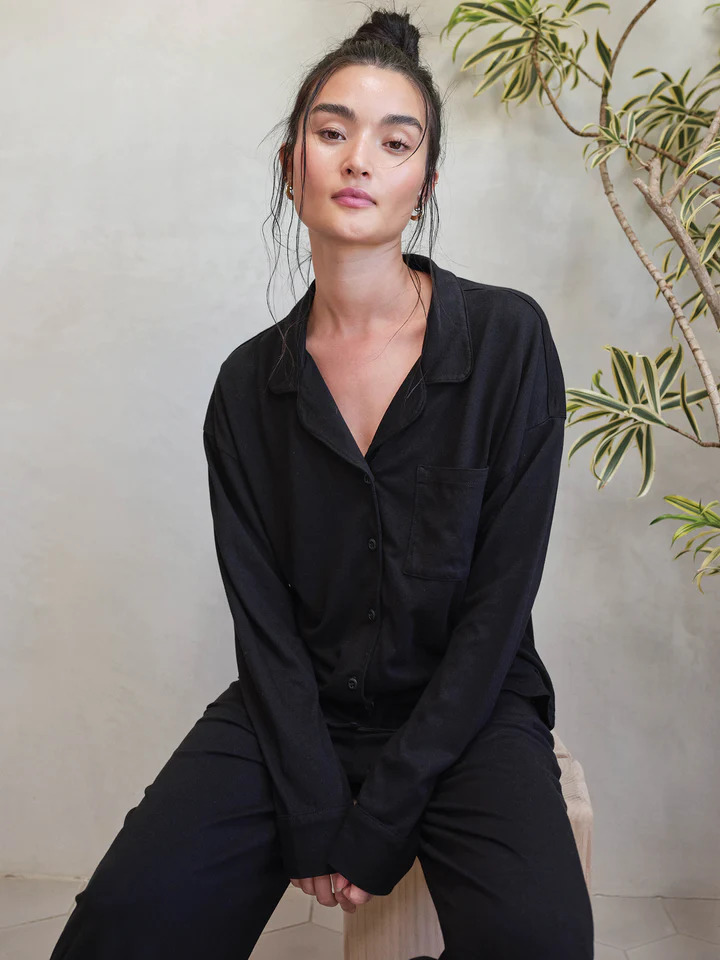 A model seated on a stool wearing a black collared long sleeve pajama set by Mate The Label.
