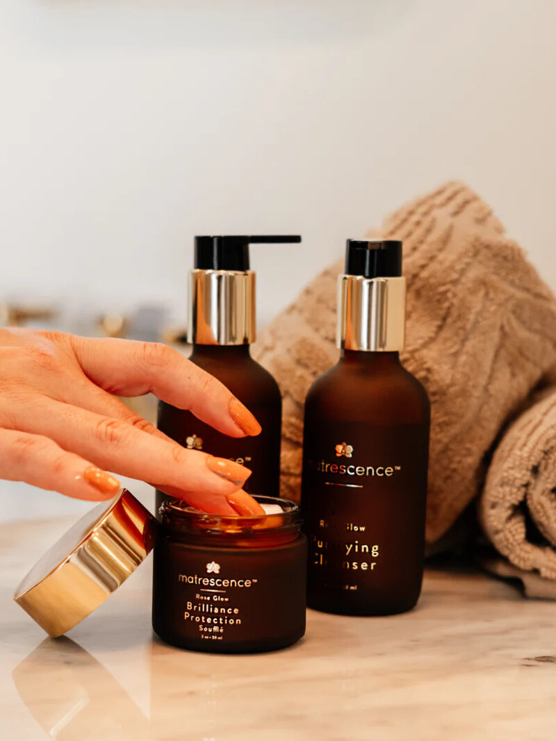 A hand dipping into a jar of Matrescence cream with other Matrescence products in the products.