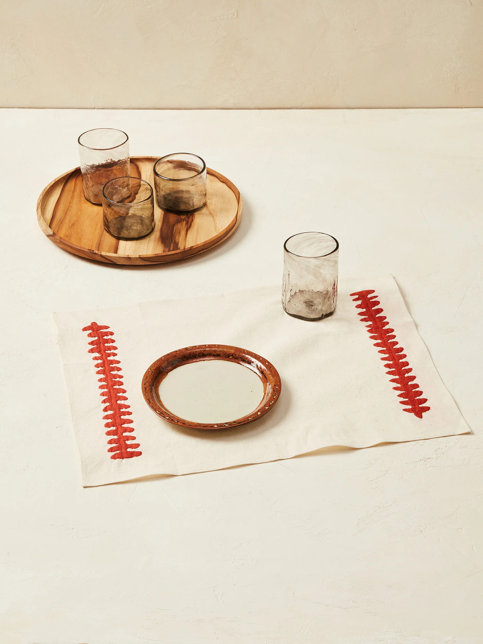 Charcoal clear glasses and a brown rimmed white plate styled on a white cloth placemat with thick red stitched details on the side. By Minna.