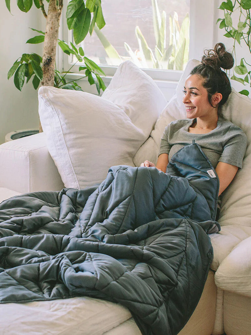 A model laying in a cream couch covered in a blue Nest Bedding Weighted Blanket. 