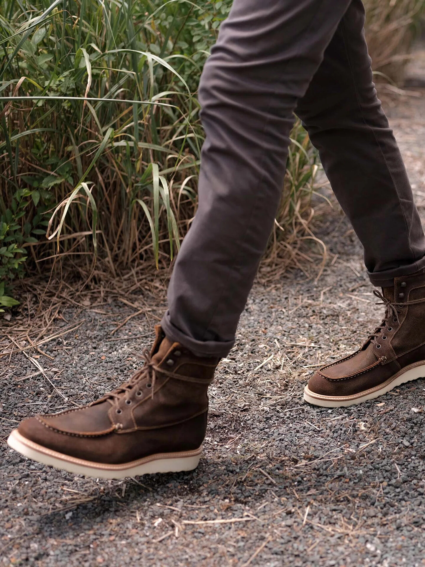 A pair of legs walking with brown-grey pants and brown Nisolo boots.