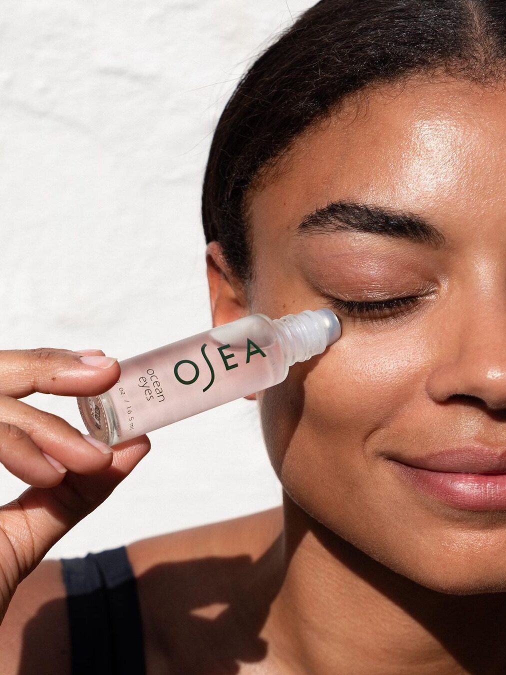 A close up of a woman applying Osea's Ocean Eyes serum to her under eye region with her eyes closed.