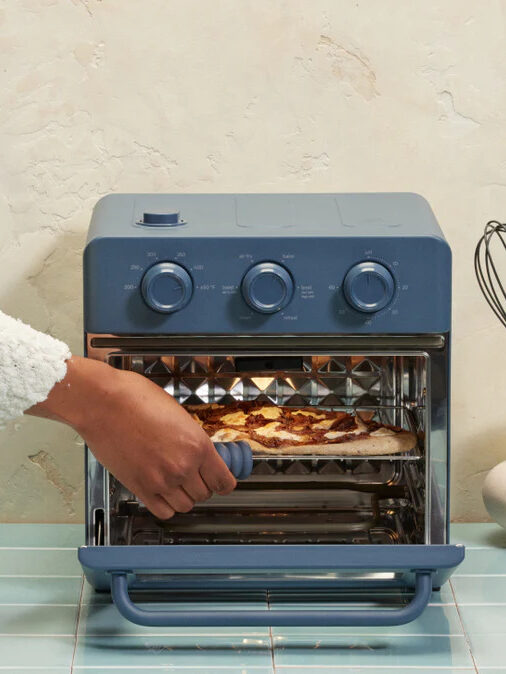 An Our Place toaster oven with the door open and a hand reaching in to remove a pizza. 