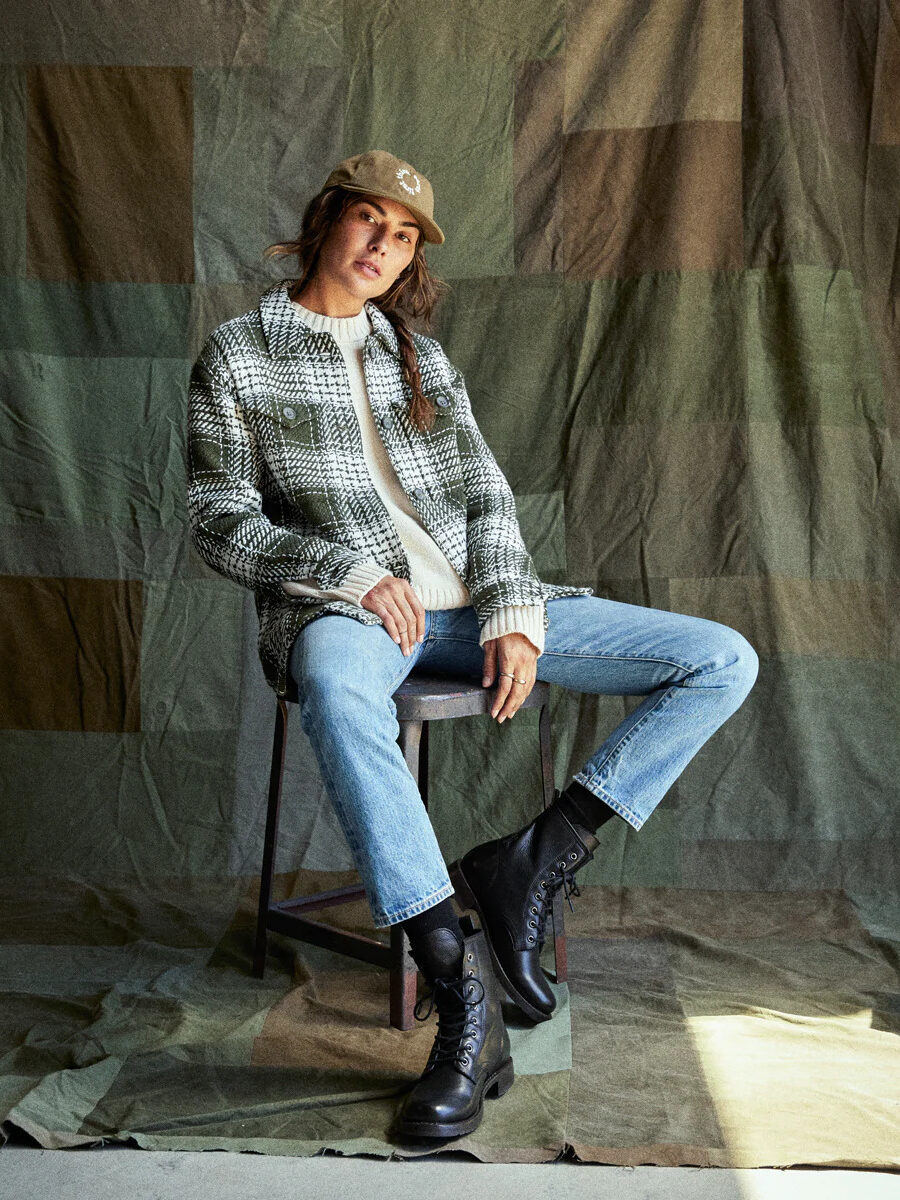 A model, seated on a stool, wearing a green and white plaid shirt jacket by Outerknown. 