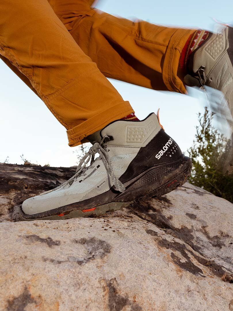 A close up of a runners feet, wearing orange pants and grey and black Salomon boots.