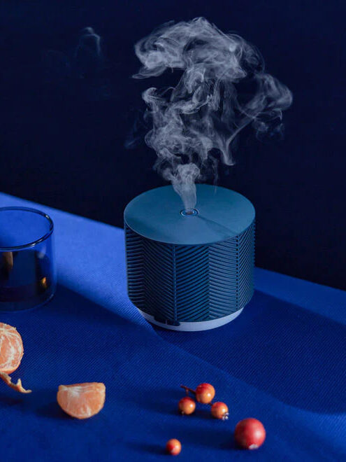 A blue Saje diffuser on a blue cloth, running with peeled oranges and cranberries scattered around.