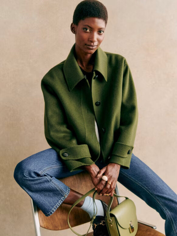 A model, seated on a wooden stool, wearing a green wool button down coat by Sezane.