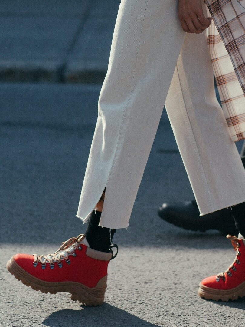 A pair of legs walking wearing white pants and red Thesus boots.
