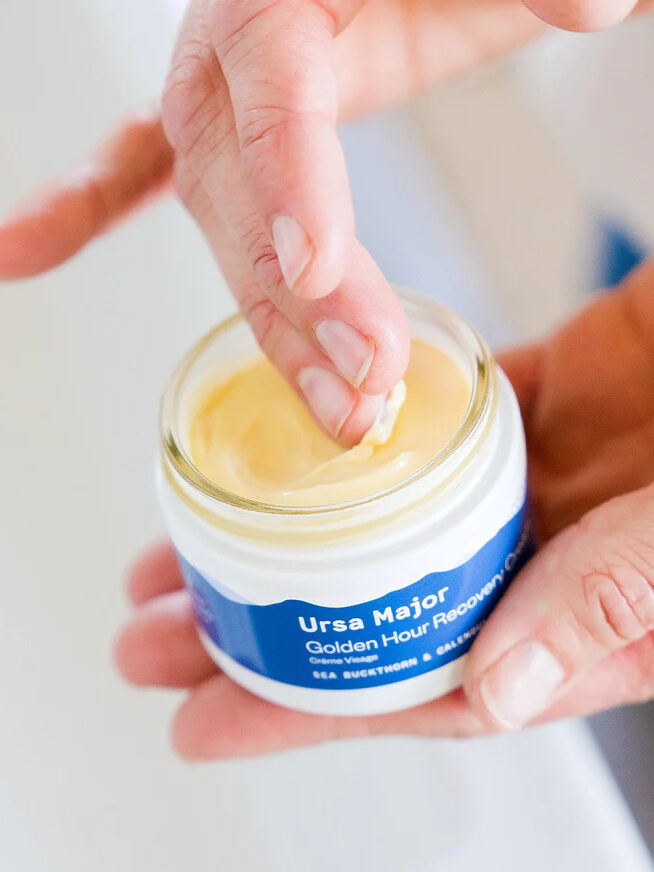 A hand scraping out some product from a jar of Ursa Major's Golden Hour Recovery Cream.