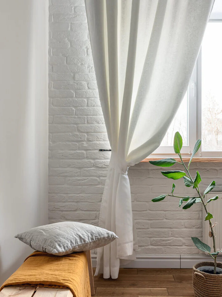 3H linen sheer white curtains gathered at the corner of a window in a room. 