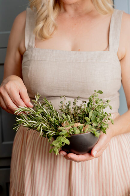 A woman in a beige dress holds a black bowl filled with fresh herbs, including rosemary, thyme, and oregano.