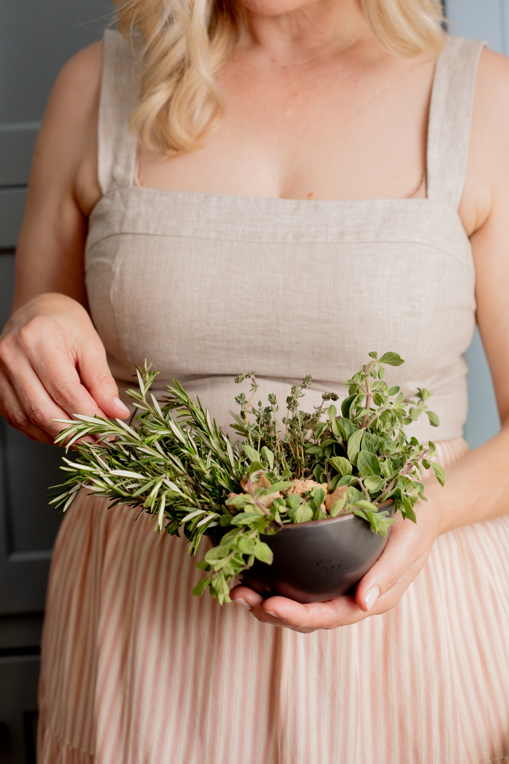 A woman in a beige dress holds a black bowl filled with fresh herbs, including rosemary, thyme, and oregano.