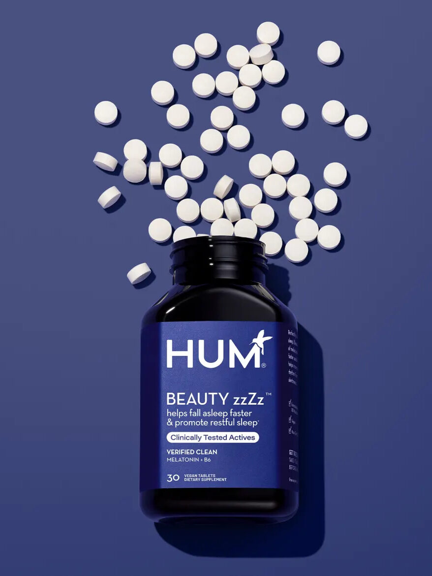 A open bottle of HUM Beauty zzZz melatonin supplements with the tablets scattered . 