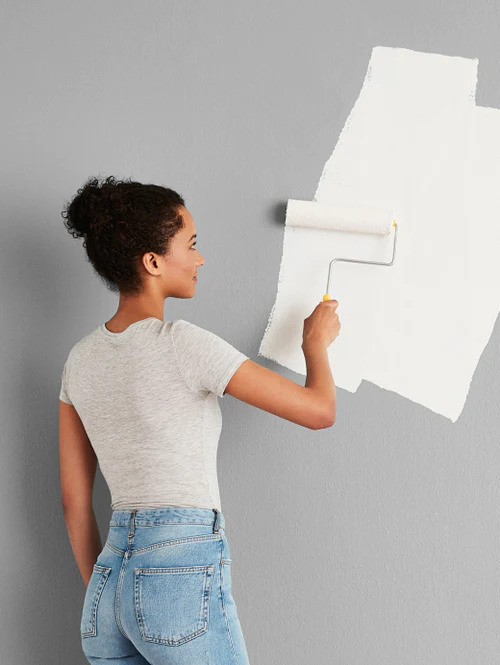 A model applying a coat of white paint on a grey wall with a roller.