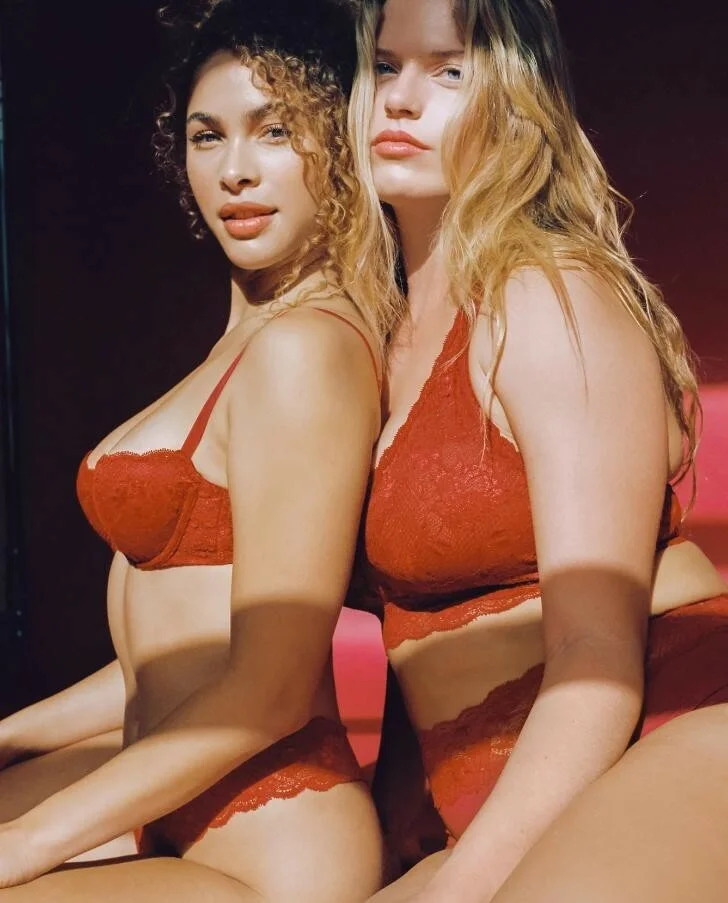 Two models sitting one behind the other, wearing Cosabella lace bras and panties in a burnt orange-red color.