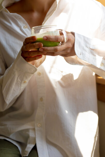 Person in a white shirt holding a glass mug with a green beverage, seated by a window with sunlight shining in.