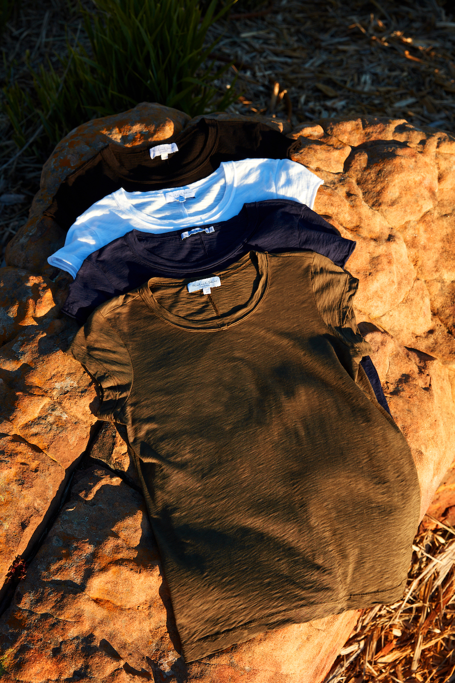 Michael Stars supima tees stacked on a rock in black, white, navy, and green.