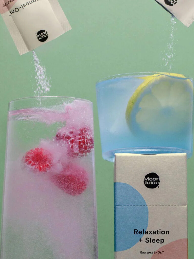 Two packs of Moon Juice's Magnesi-om being poured into glass cups, one with a pink hue and one with a blue hue. 