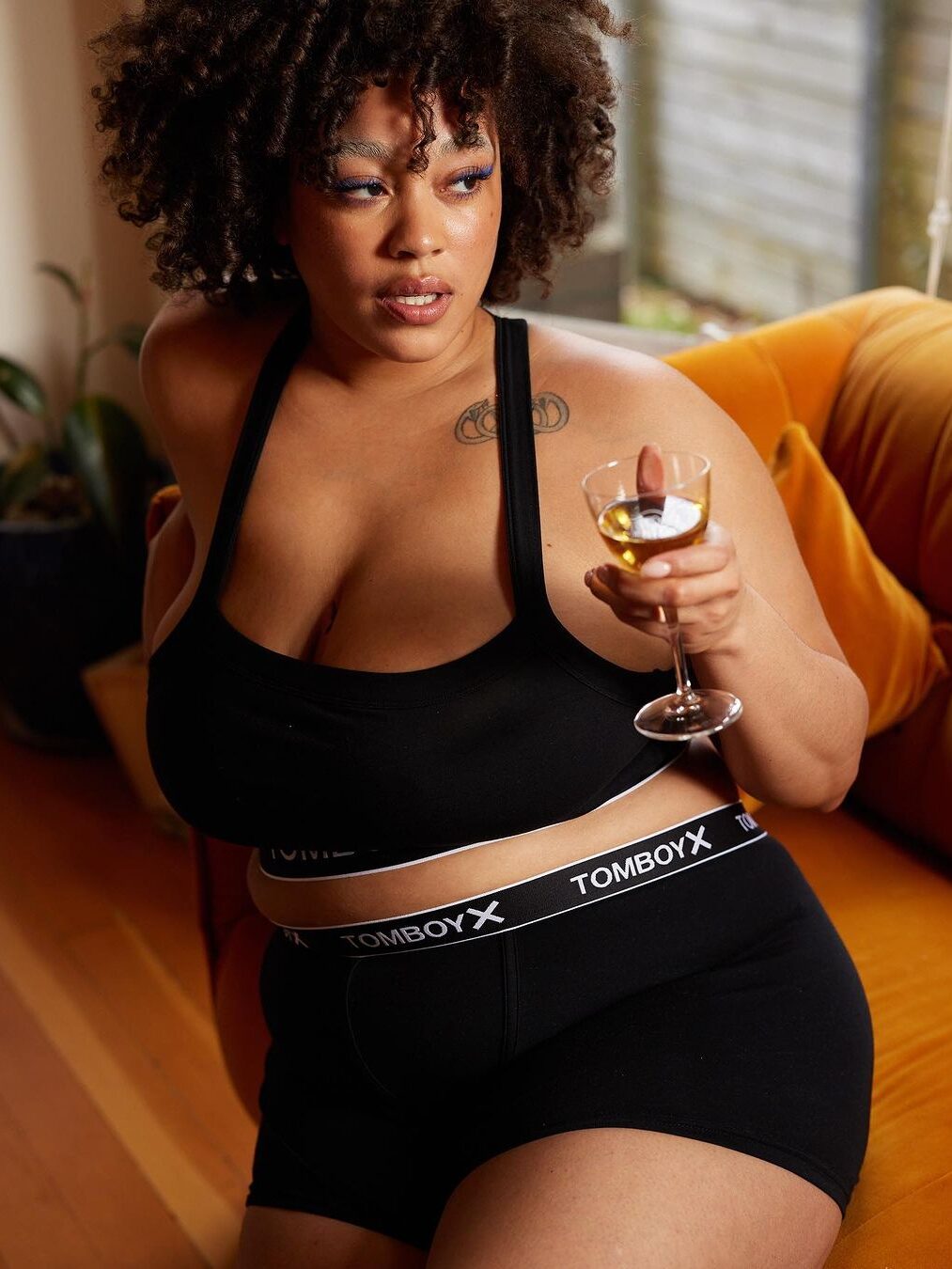 A model seated in a bedroom setting holding a glass of white wine, wearing a black TomboyX lingerie set, including boyshorts and a bralette.