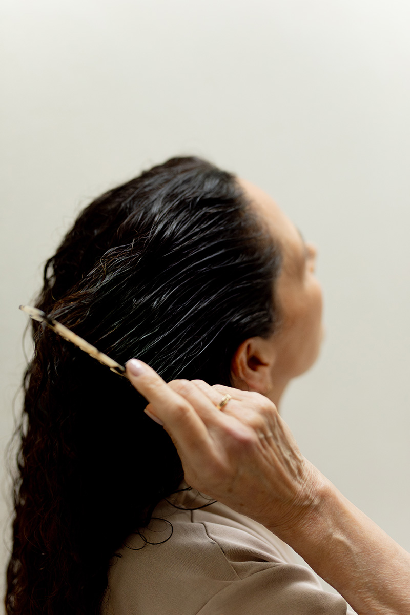 A woman combs her long, dark hair with a wooden comb. she is in profile view, focusing on her hair against a light background.