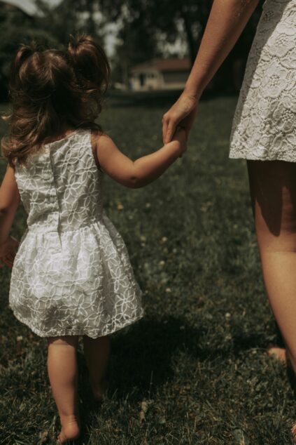 A young child and an adult, both wearing white lace dresses, hold hands while walking on grass outdoors.