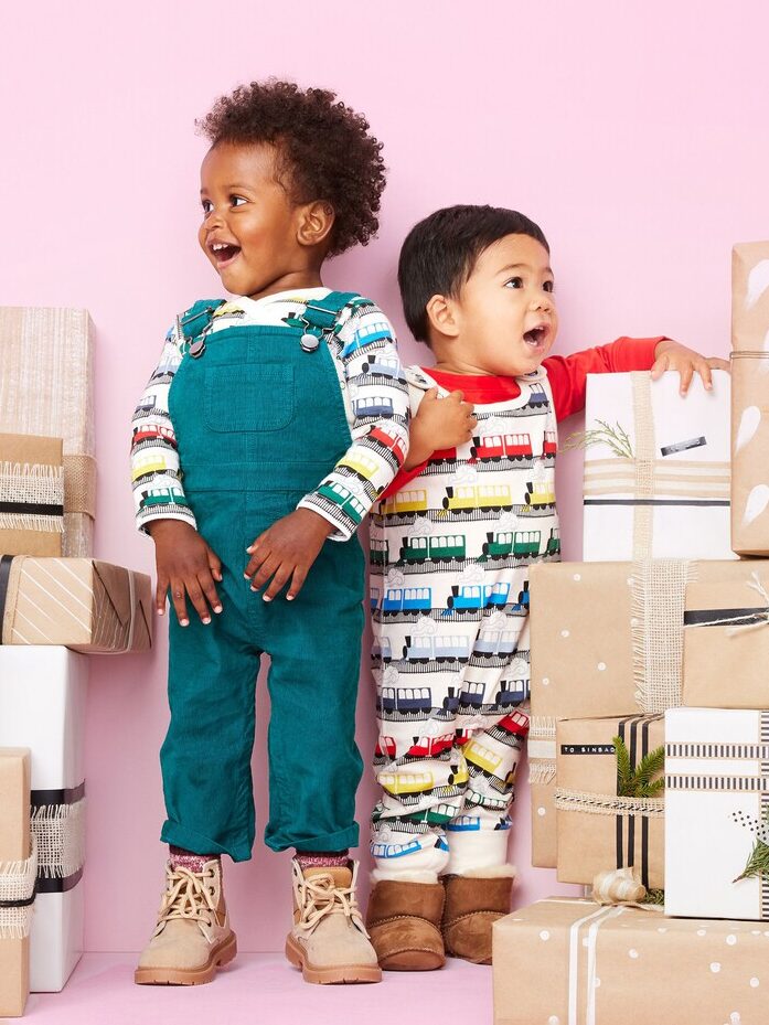 Two babies wearing matching train patterned onesies from Hanna Andersson, with one wearing teal overalls on top.