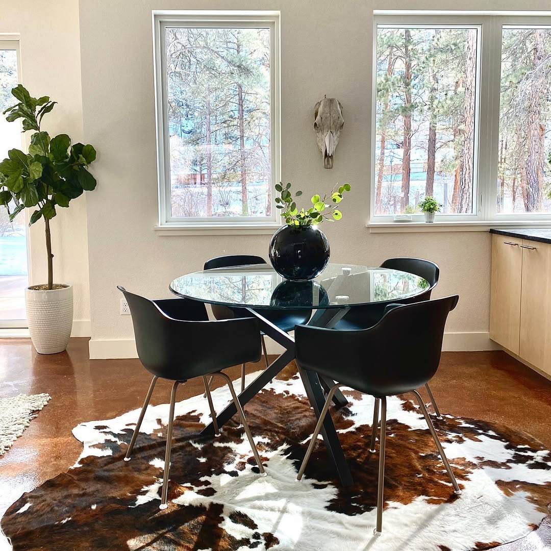 A modern dining area with a round glass table, four black chairs, a black vase with greenery, a cowhide rug, and large windows overlooking trees.