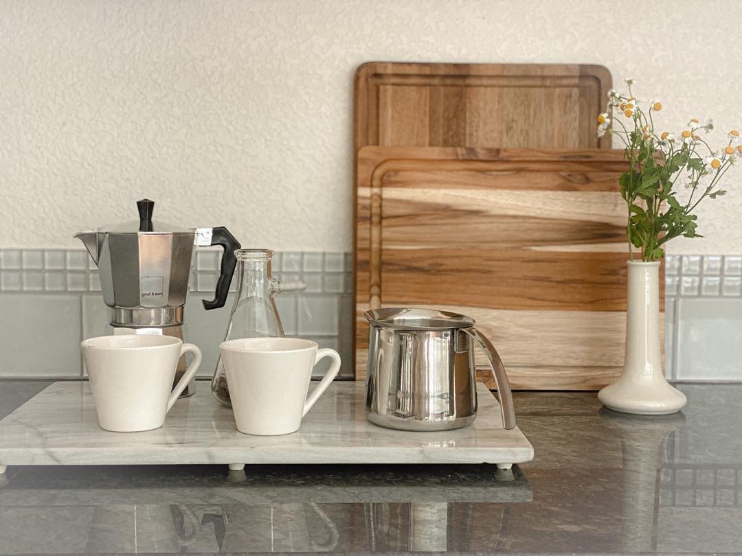 Two coffee cups, a moka pot, a glass carafe, and a metal milk frother are on a marble tray; wooden cutting boards and a vase with flowers are in the background.