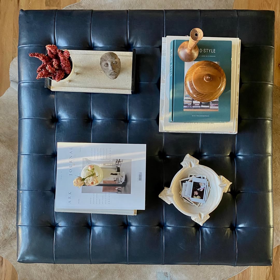 A black tufted leather ottoman with books, a tray holding red dried flowers and a face sculpture, a wooden vessel, and a dish holding a small photo.