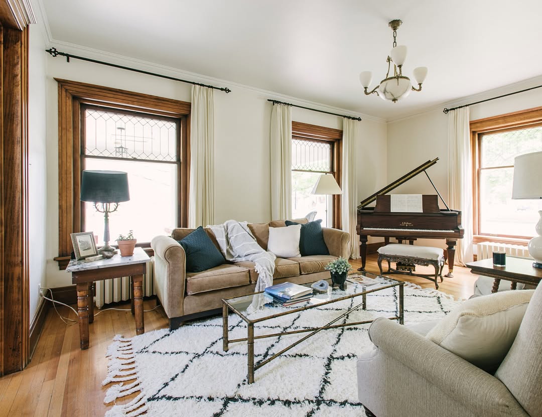 Living room with beige sofas, glass coffee table, patterned rug, two windows with curtains, table lamps, and a grand piano in the corner.
