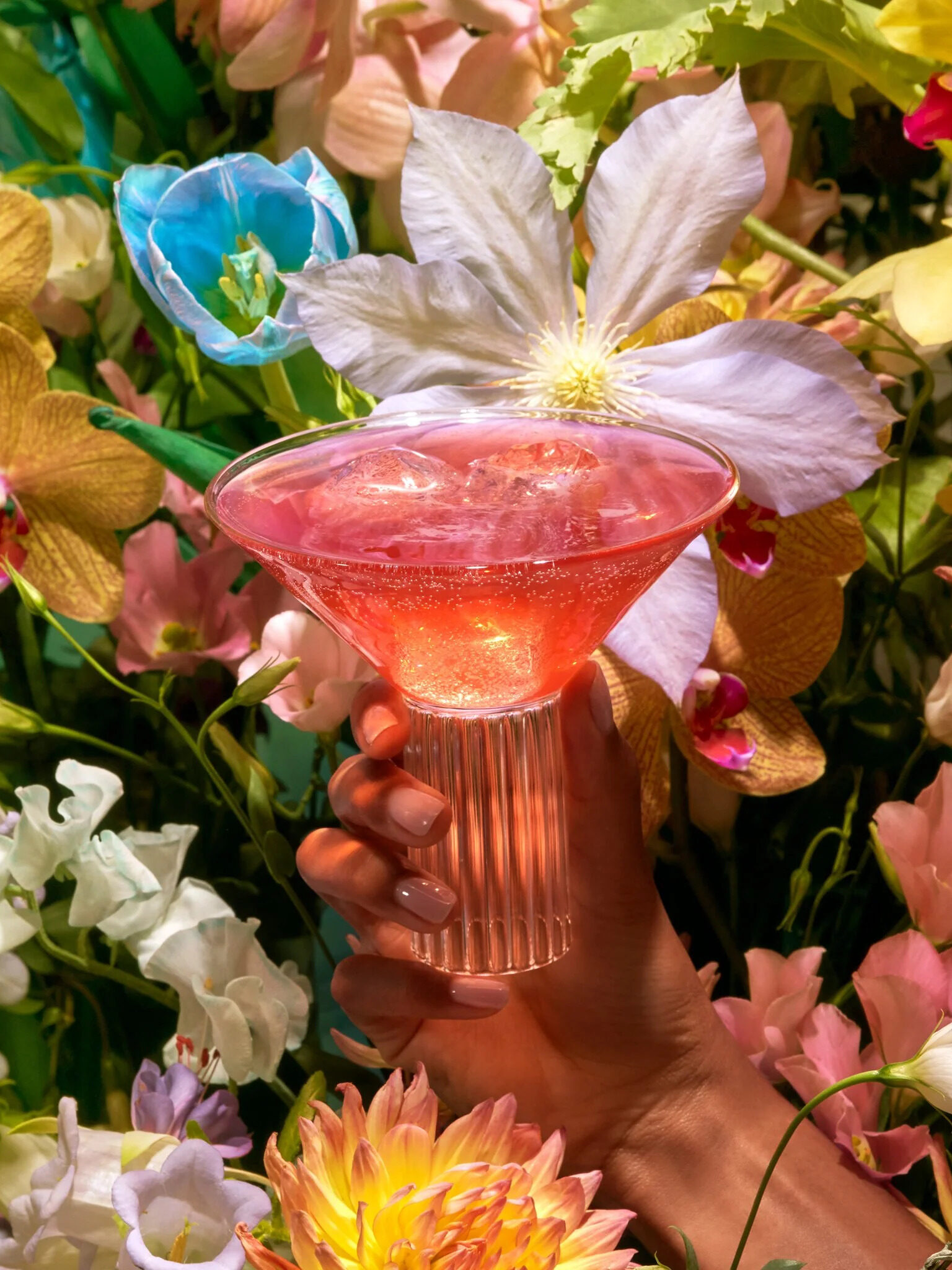 A hand holding up a cocktail glass filled with Kin Euphorics, with flowers in the background.