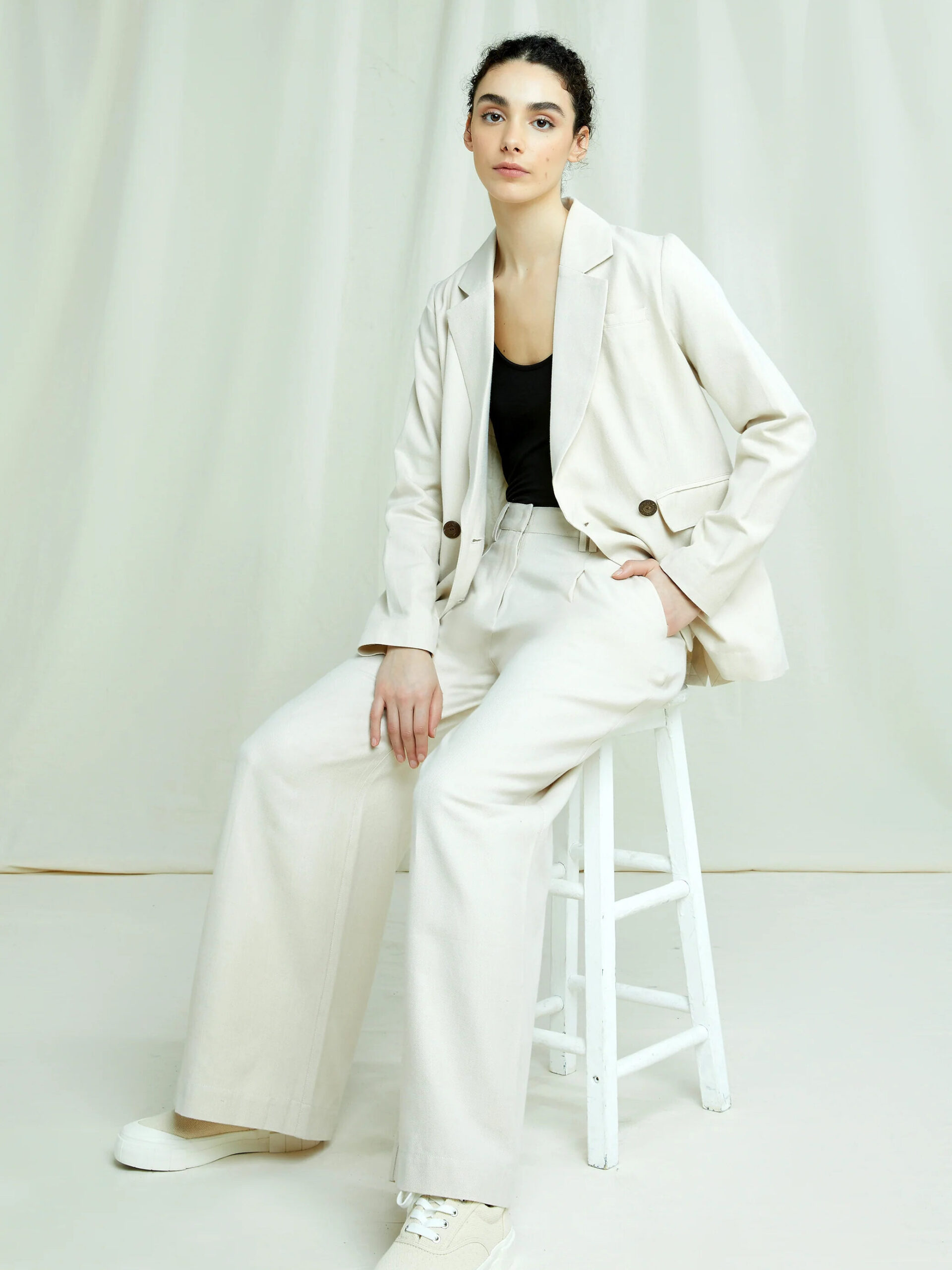 A model seated wearing People Tree's Naira Blazer and matching pants in cream.