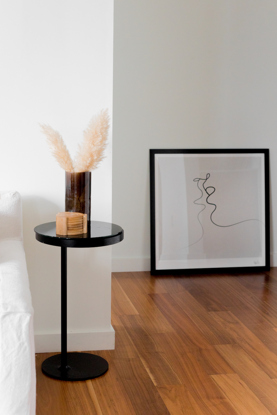 Minimal bedroom scene with a sliver of white bedding, a bedside table with a vase of dried grass, and a frame laid on the floor, resting on the wall