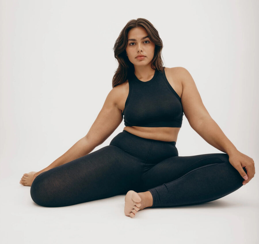 A studio shot of a model seated in pigeon pose wearing a black Organic Basics cotton leggings and high neck sports bra set.